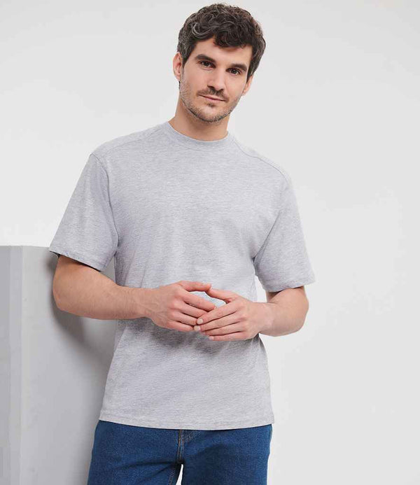 A man with short dark hair and a trimmed beard stands indoors against a white background, wearing the Russell Athletic Heavyweight T-Shirt in light gray with a ribbed neck trim by Russell Athletic and blue jeans, his hands loosely clasped.