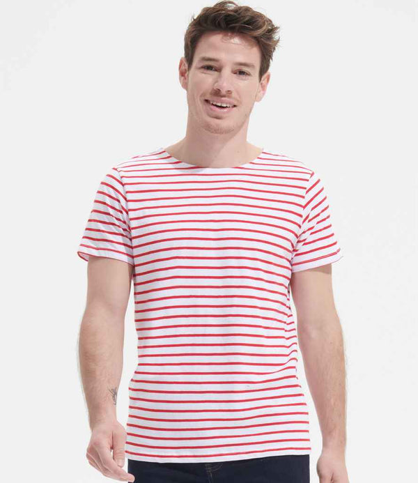 A man faces forward against a white background, smiling slightly, wearing the SOLS Miles Striped T-Shirt by SOLS. The fitted white tee features horizontal red stripes, a self fabric neckline for comfort, and is paired with dark pants.