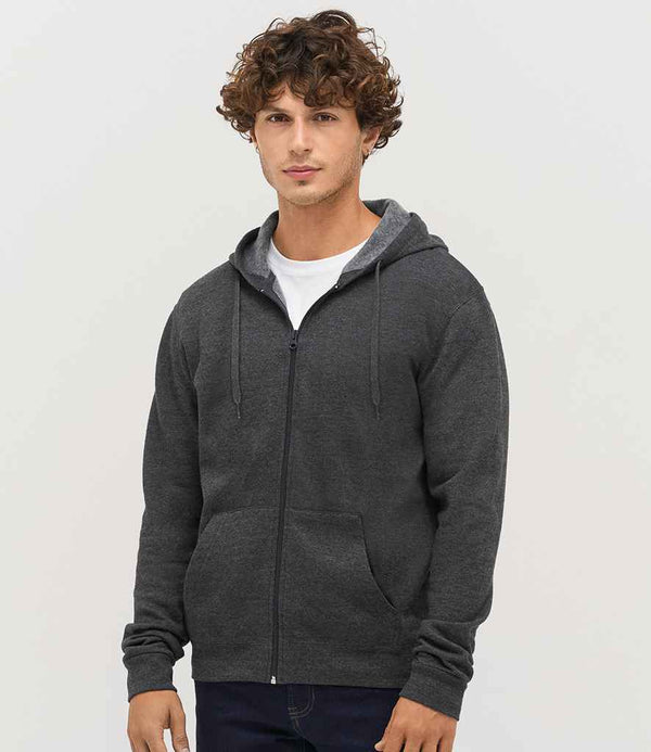 A young man with curly brown hair wears the SOLS Stone Zip Hooded Sweatshirt by SOLS in dark gray over a white t-shirt and dark pants, standing against a plain light background and looking at the camera with a neutral expression.