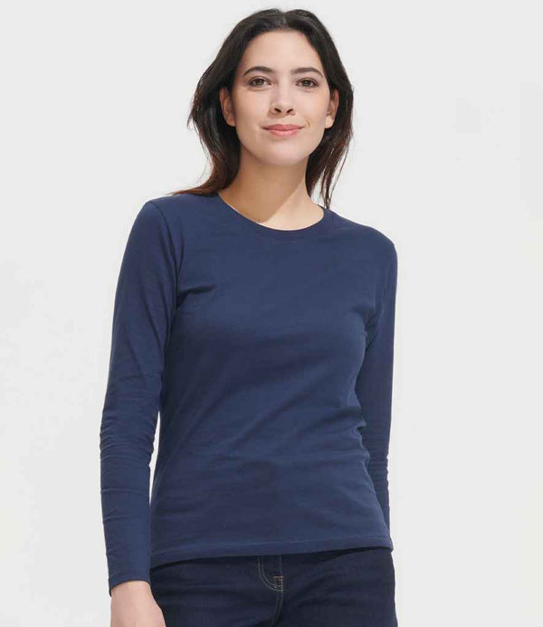 A person with long dark hair is standing and wearing the SOLS Ladies Imperial Long Sleeve T-Shirt by SOLS in navy blue and dark jeans, facing the camera with a neutral expression against a plain white background.