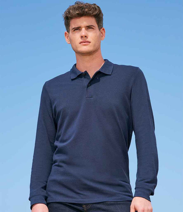A man stands outdoors under a clear blue sky, wearing the SOLS Perfect Long Sleeve Piqué Polo Shirt in navy with a ribbed collar and two-button placket by SOLS, paired with dark jeans. He faces forward with a neutral expression.