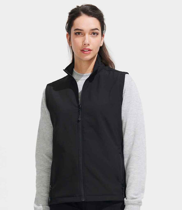 A woman with long dark hair is wearing the SOLS Ladies Race Soft Shell Bodywarmer by SOLS over a light gray long-sleeve shirt, standing against a plain light gray background and facing forward with a neutral expression.