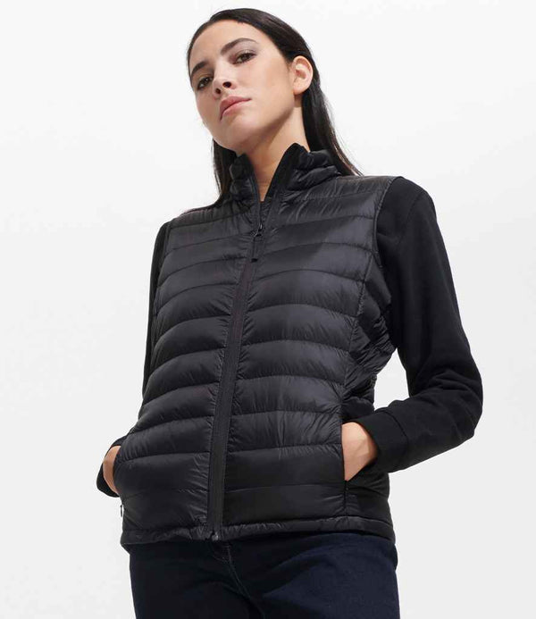 A woman stands with her hands in the pockets of a SOLS Ladies Wilson Lightweight Padded Bodywarmer, worn over a black long-sleeve shirt. She looks slightly down with a neutral expression against a plain white background.