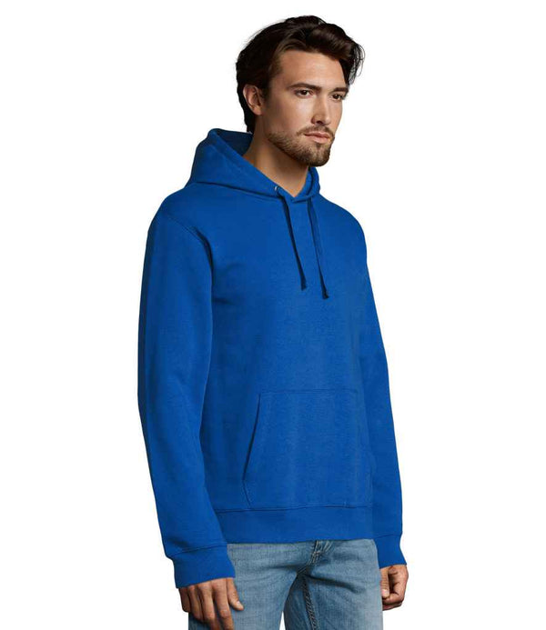 A man with brown hair and a trimmed beard wears the SOLS Unisex Spencer Hooded Sweatshirt in bright blue, featuring a front pouch pocket, paired with light blue jeans, standing against a plain white background.