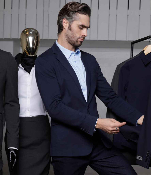 A man in a navy suit examines the sleeve of a NEOBLU Marcel Piqué Blazer on a rack. Next to him stands a mannequin with a gold reflective head dressed in formal wear, with other NEOBLU suits visible in the background.