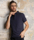 A man with dark hair and a beard wears the NEOBLU Owen Piqué Polo Shirt with a ribbed collar, standing against a light textured wall. He looks slightly to the side, one hand touching his chin. Brand: NEOBLU.