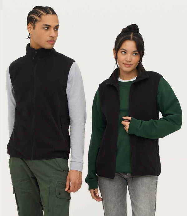 A man and a woman wear SOLS Unisex Factor Recycled Micro Fleece Bodywarmers in black, featuring full zip and front zip pockets. He pairs his with a light grey shirt and green cargo pants; she wears a dark green sweatshirt and light grey jeans.