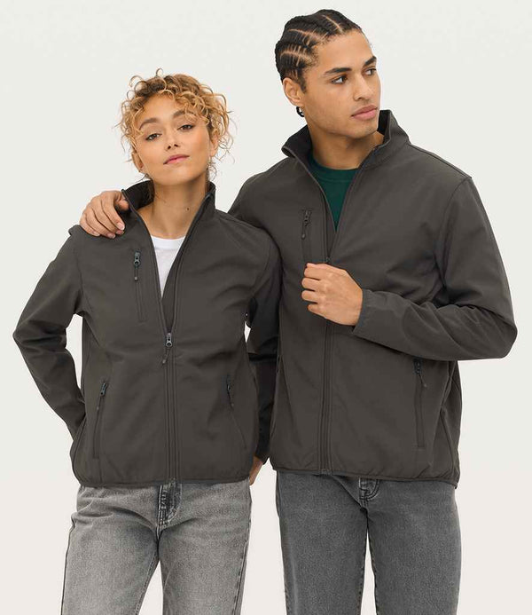 Two people stand side by side wearing SOLS Ladies Falcon Recycled Soft Shell Bodywarmers in dark gray and light gray jeans. One rests an arm on the others shoulder as they look to the side with neutral expressions.
