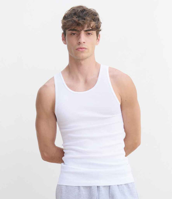 A young man with short, curly brown hair models the SOLS Pablo Ribbed Tank Top in white and light gray pants, posing against a plain, light background with his arms behind his back.