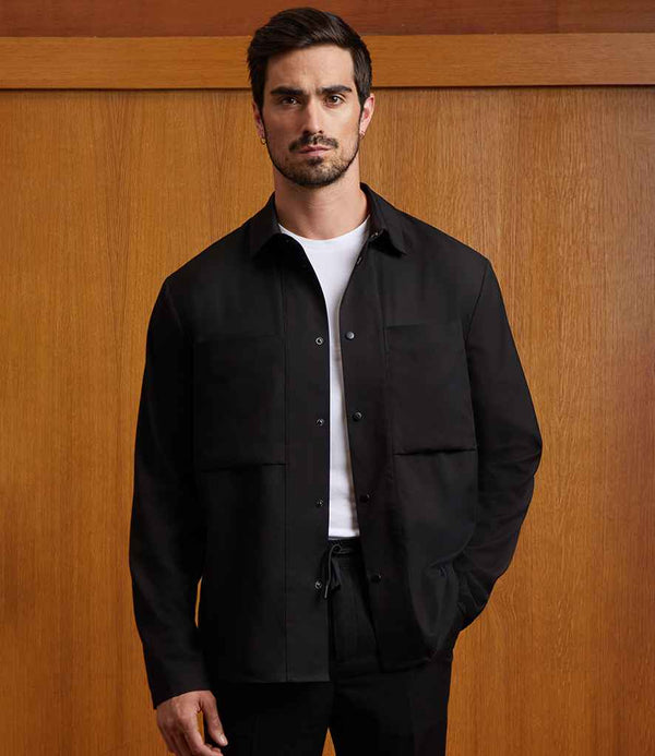A man with short dark hair and a beard stands before a wooden wall, wearing the NEOBLU Unisex Madden Overshirt over a white t-shirt and black pants, one hand in his pocket as he looks straight ahead.