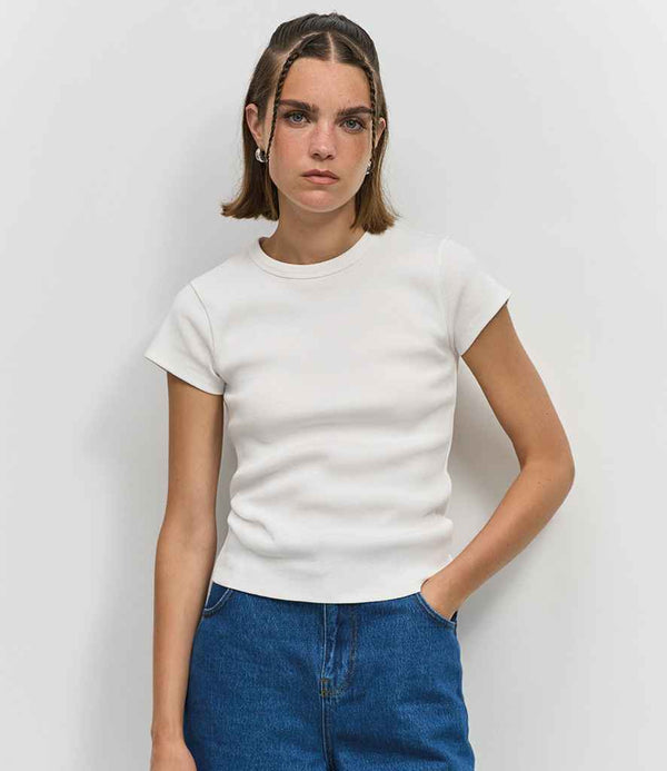 A young woman with straight brown hair and two thin braids stands against a white wall, wearing the SOLS Ladies Sara T-Shirt by SOLS in a fitted short-sleeve white style, paired with blue jeans and a neutral expression.