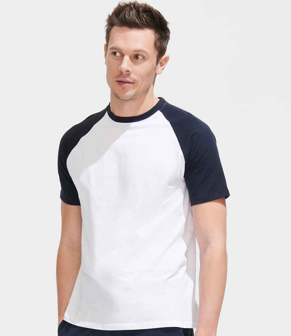 A man stands against a plain light gray background, looking slightly to the side, wearing the SOLS Funky Contrast Baseball T-Shirt by SOLS with short sleeves, contrast raglan sleeves, and a ribbed collar.