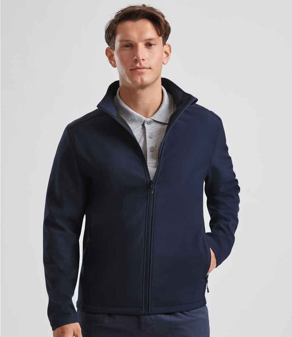 A man with short brown hair and a neutral expression faces forward in a plain, light background, wearing a Russell Athletic Essential Recycled Soft Shell Jacket in navy over a grey collared shirt. The jacket is made from recycled polyester.