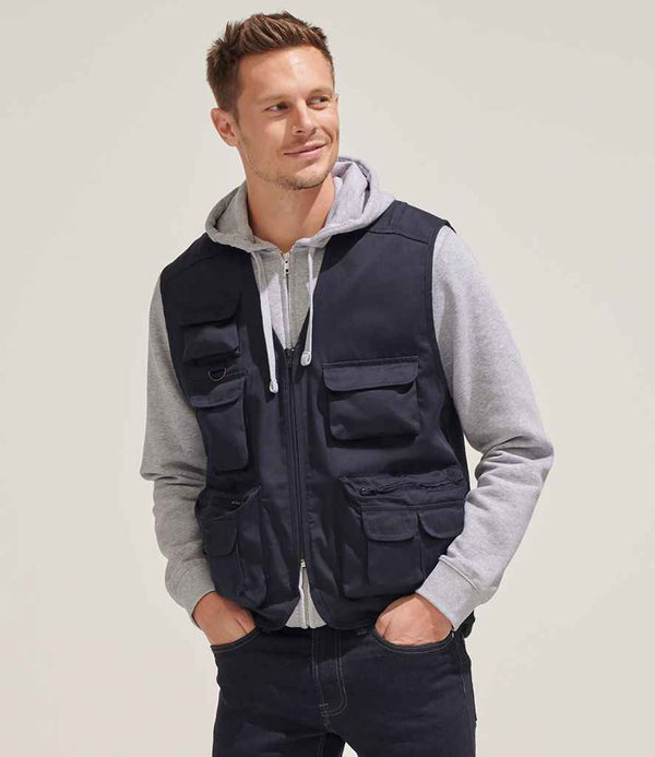 A man in dark jeans and a grey hooded sweatshirt layers the SOLS Wild Waistcoat by SOLS—featuring nine pockets, including a phone pocket and mesh-lined back—and stands with hands in his pockets, looking slightly to the side.