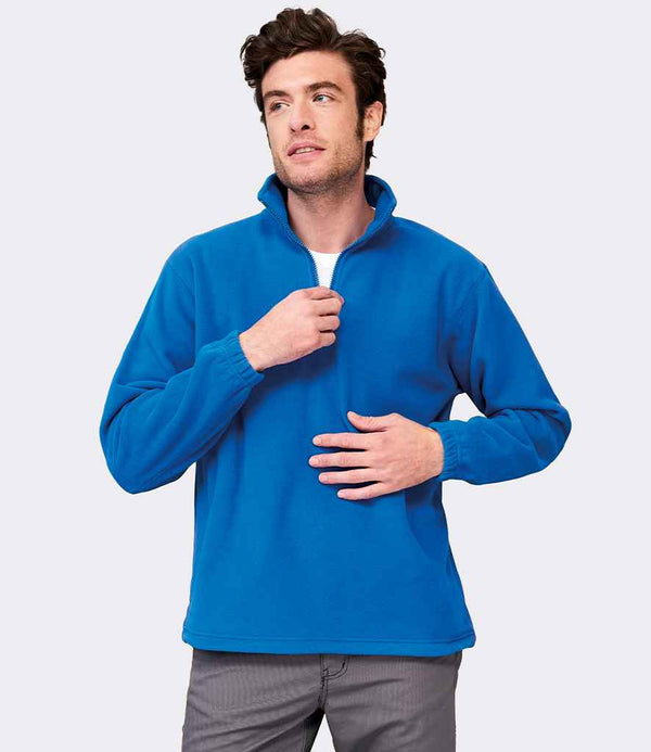 A man wears the SOLS Ness Zip Neck Fleece by SOLS in royal blue, featuring a quarter-zip collar and elasticated cuffs, standing against a plain light grey background while partially zipping up the high-density fleece.