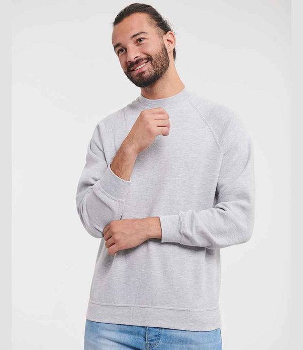 A bearded man with tied-back hair wears the Russell Athletic Raglan Sweatshirt by Russell Athletic, featuring a ribbed collar and fade resistant light grey fabric, paired with light blue jeans. He stands against a white background smiling with one hand raised.