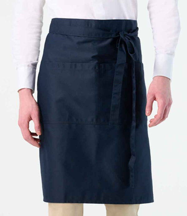 A person in a white shirt and beige pants is shown from the shoulders down, modeling the SOLS Greenwich Apron by SOLS—a dark blue apron with large front pocket and self-fabric ties, both practical and stylish for everyday use.