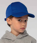 A young child in a SOLS Kids Sunny Cap by SOLS, featuring a bright blue 5 panel design with ventilation eyelets, and a light gray hoodie, looks slightly to the side against a plain background.