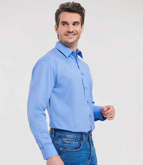 A man in the Russell Athletic Collection Long Sleeve Easy Care Poplin Shirt—light blue with adjustable cuffs and a classic collar—pairs it with blue jeans and stands against a white background, looking left with a slight smile.
