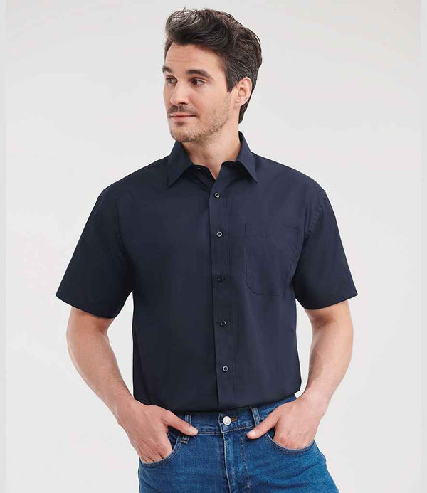 A man stands against a plain light background, wearing the Russell Athletic Collection Short Sleeve Easy Care Poplin Shirt in dark navy with a classic collar and chest pocket, paired with blue jeans. He looks slightly right with his hands in his pockets.
