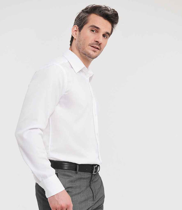 A man wearing the Russell Athletic Collection Long Sleeve Tailored Ultimate Non-Iron Shirt in white, paired with dark grey trousers and a black belt, stands against a plain light background facing slightly to the right.