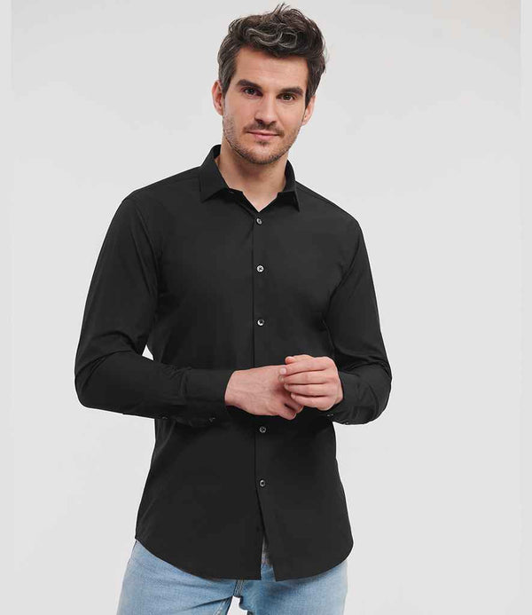 A man wears the Russell Athletic Collection Long Sleeve Ultimate Stretch Shirt in black with a semi-cut away collar and light blue jeans, standing against a white background and looking at the camera with a neutral expression.