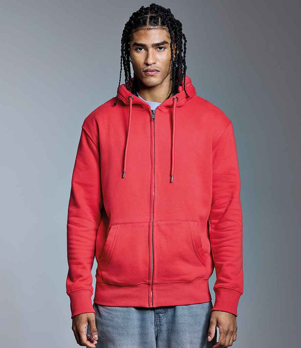 A person with long braided hair wears the Anthem Organic Full Zip Hoodie by Anthem— a slim fit, cotton faced hoodie with hood and drawstrings—paired with light blue jeans, standing against a plain gray background with a neutral expression.