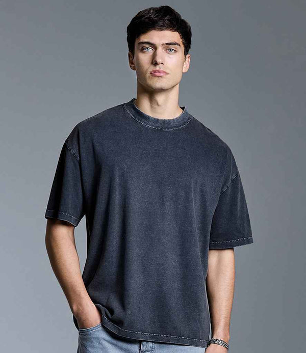 A man with short dark hair wears the Anthem Distortion Washed T-Shirt by Anthem and light blue jeans, standing against a plain gray background with his left hand in his pocket.