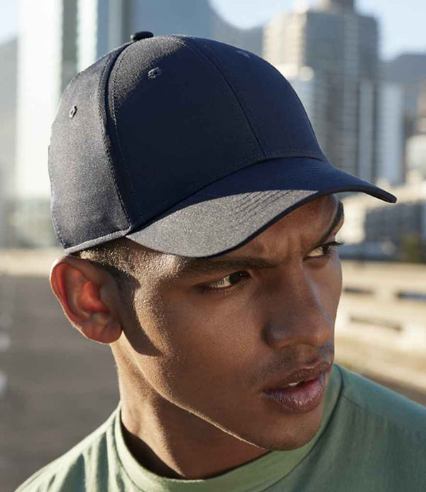 A person in a green shirt wears the Beechfield Performance Cap by Beechfield, a dark blue lightweight and breathable cap, while looking to the side with blurred city buildings and outdoor sunlight in the background.