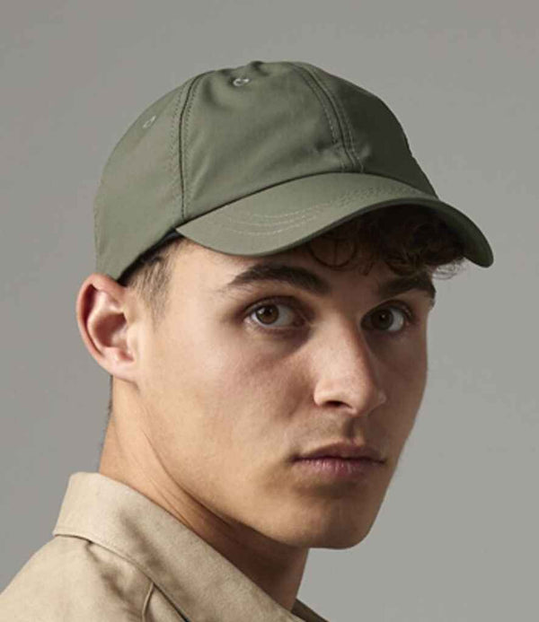 A young person with short curly hair wears the Beechfield Outdoor 6 Panel Cap in olive green by Beechfield and a beige shirt, looking slightly past the camera against a plain background.