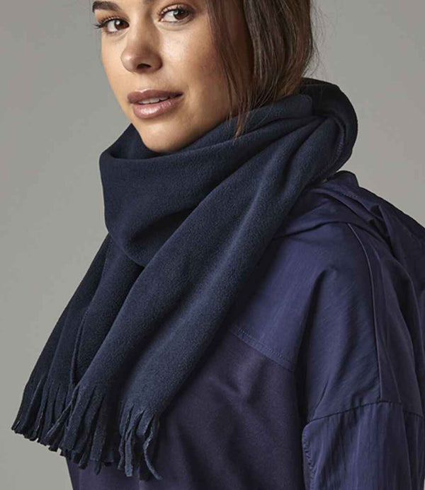 A woman wearing the Beechfield Suprafleece® Dolomite Scarf by Beechfield in dark blue and a matching top looks to the side with a neutral expression, posed against a gray background. The scarf provides lightweight warmth for effortless comfort.