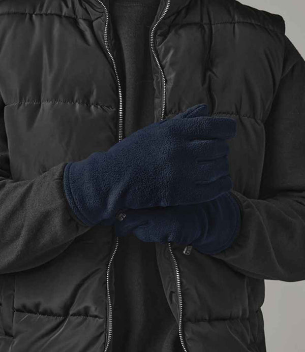 Wearing a black puffer vest, black long-sleeve shirt, and Beechfield Recycled Fleece Gloves, someone stands with arms crossed against a neutral background, highlighting the lightweight warmth and comfort of Beechfield’s eco-friendly gloves.