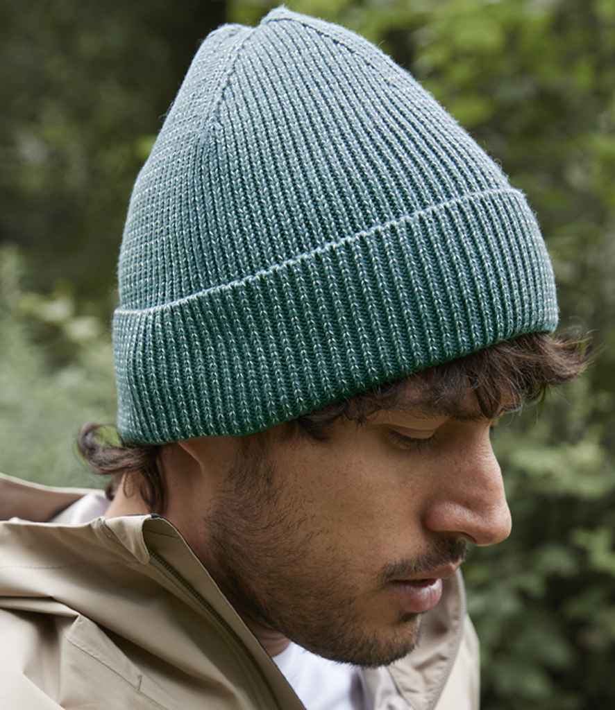 A man wearing the Beechfield Streetlite Reflective Beanie in teal by Beechfield and a beige jacket looks down outdoors, with green foliage in the background.