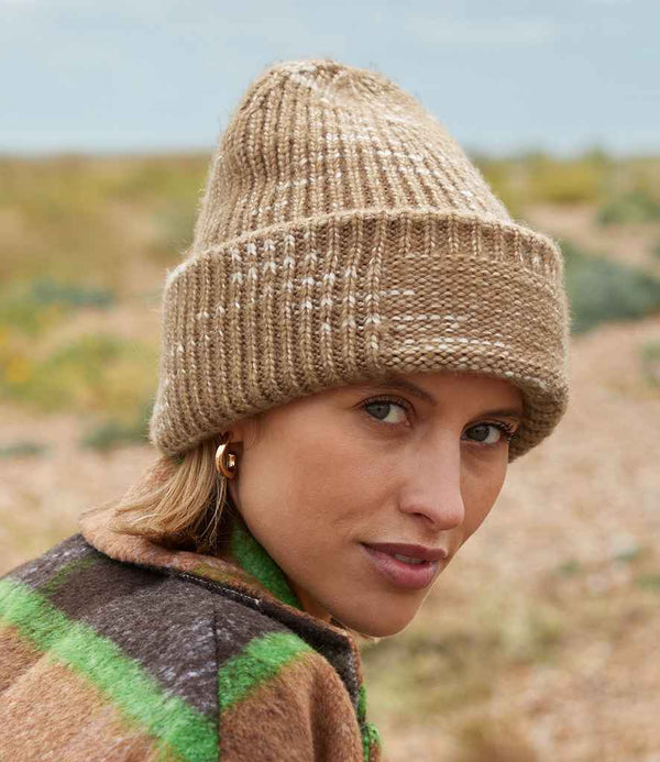A person wearing a Beechfield Chunky Knitted Patch Beanie in tan and a brown plaid jacket with green stripes stands outdoors on rocky terrain, looking at the camera.