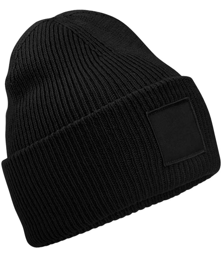 The Beechfield Deep Cuff Tonal Patch Beanie by Beechfield features a plain black ribbed knit, a wide folded brim, and a blank tonal rectangular patch on the front. Shown on a white background.