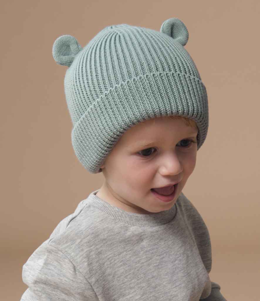 A young child wears a Beechfield Organic Baby Bear Beanie, a light blue ribbed knit hat with 3D ear detail by Beechfield, paired with a light gray sweater, looking slightly to the side against a plain beige background.
