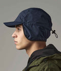 In profile, a person wears the Beechfield Mountain Cap by Beechfield—a dark blue waterproof hat with fold-down earflaps and adjustable drawstring—paired with a green jacket and black hood. The background is plain and neutral.