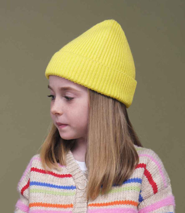 A young girl with straight, shoulder-length blonde hair wears the Beechfield Kids Fashion Beanie by Beechfield in yellow and a cream sweater with colorful stripes, looking left against a plain olive green background.