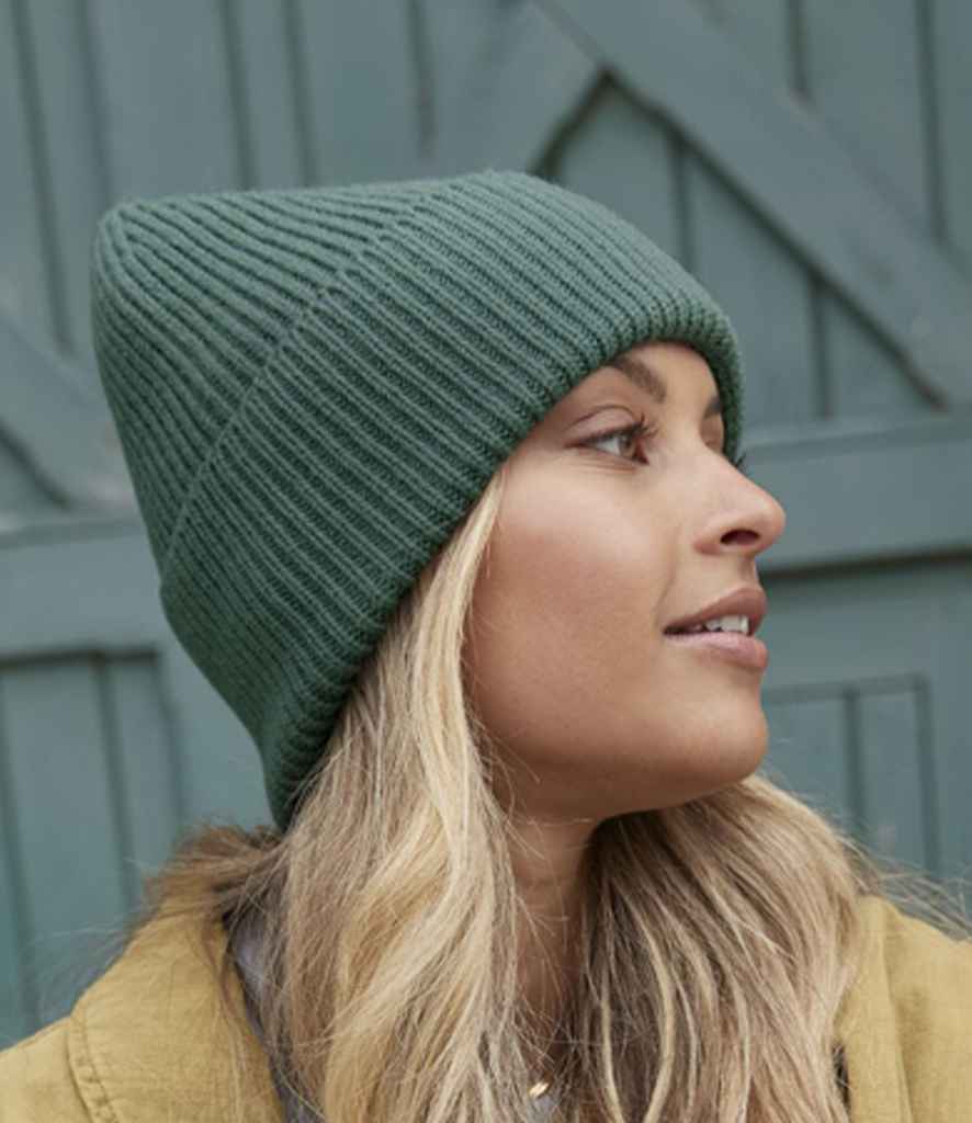A person with long blonde hair wears a Beechfield Recycled Oversized Cuffed Beanie in green and a tan jacket, looking right, with a wooden door in the background.