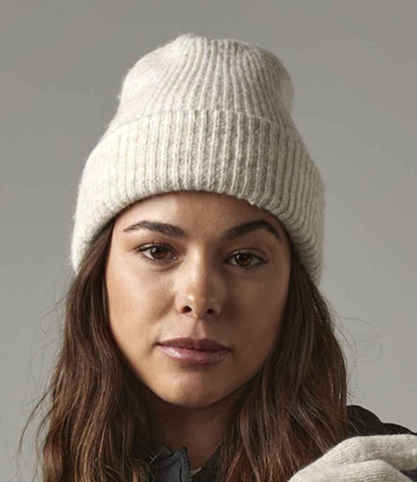 A person with long brown hair wears a light-colored Beechfield Cosy Ribbed Beanie by Beechfield and a dark jacket, looking at the camera against a plain gray background.