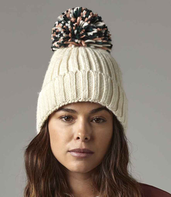 A person with long brown hair wears the cream-colored Beechfield Hygge Beanie by Beechfield, featuring a ribbed knit design and a large luxury pom pom, set against a plain gray background.
