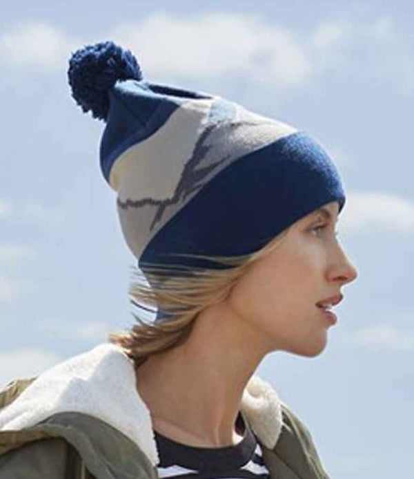 A person in profile wears the Beechfield Mountain Peaks Pom Pom Beanie in blue and gray, paired with a striped shirt and shearling-collared jacket. The sky is visible in the background.