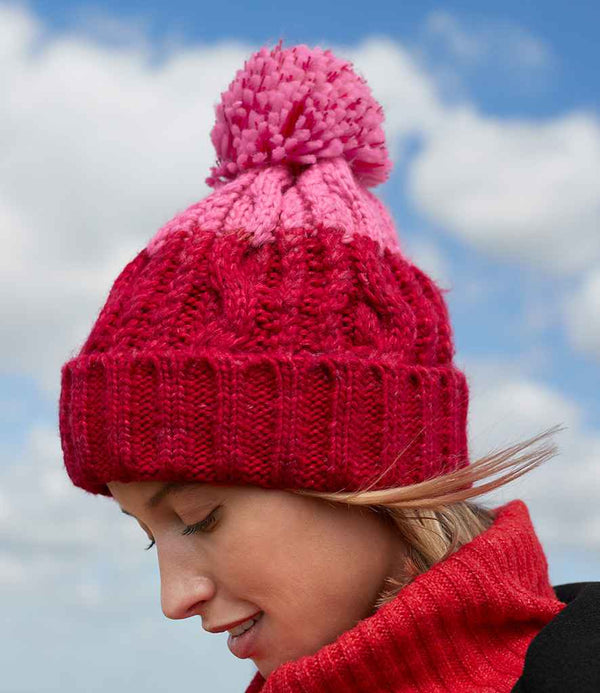 A person wearing the Beechfield Apres Beanie in red and pink with a pom-pom and a red turtleneck sweater looks downward against a partly cloudy sky. The beanie is by the brand Beechfield.