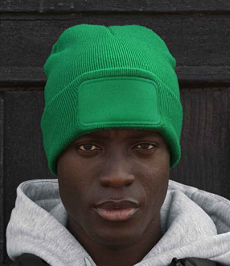 A person in a green Beechfield Original Patch Beanie featuring a cotton twill patch and a light gray hoodie stands before a dark wooden background.