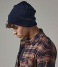 A person wearing a Beechfield Thinsulate™ Beanie in navy blue, featuring a double-layer knit and non-ribbed turn up, with a red and beige plaid shirt, looks downward against a plain grey background.