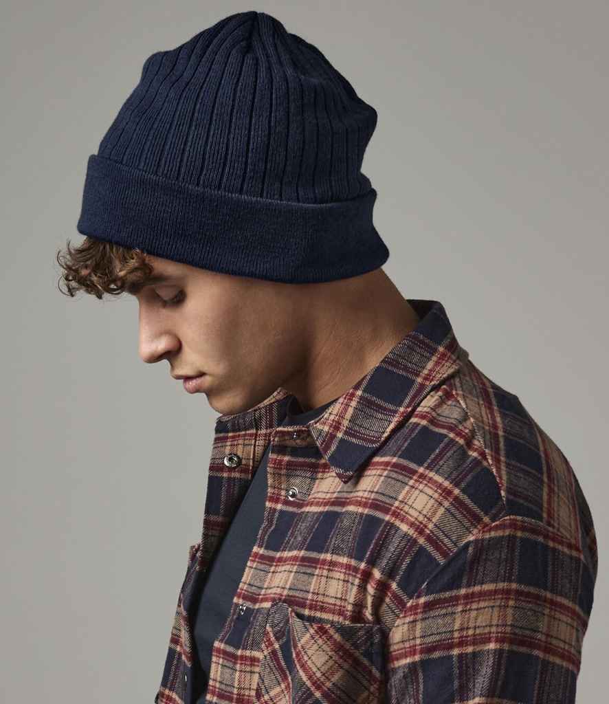 A person wearing a Beechfield Thinsulate™ Beanie in navy blue, featuring a double-layer knit and non-ribbed turn up, with a red and beige plaid shirt, looks downward against a plain grey background.