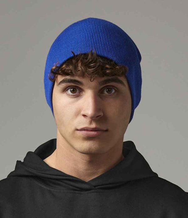 A young person with curly brown hair wears a Beechfield Recycled Original Pull-On Beanie in blue and a black hoodie, looking directly at the camera with a neutral expression against a plain gray background.