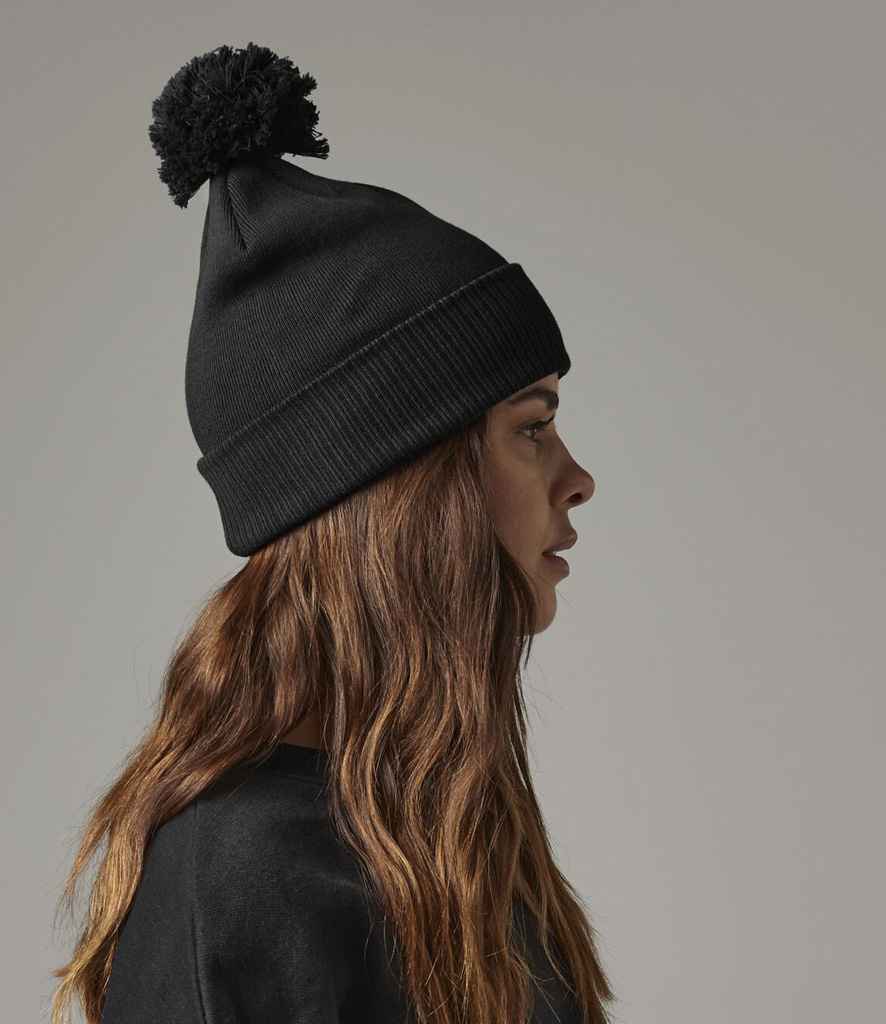 A person with long brown hair, shown in profile against a plain light gray background, wears the Beechfield Organic Cotton Snowstar® Beanie by Beechfield—a double-layer knit black beanie featuring a pom-pom.