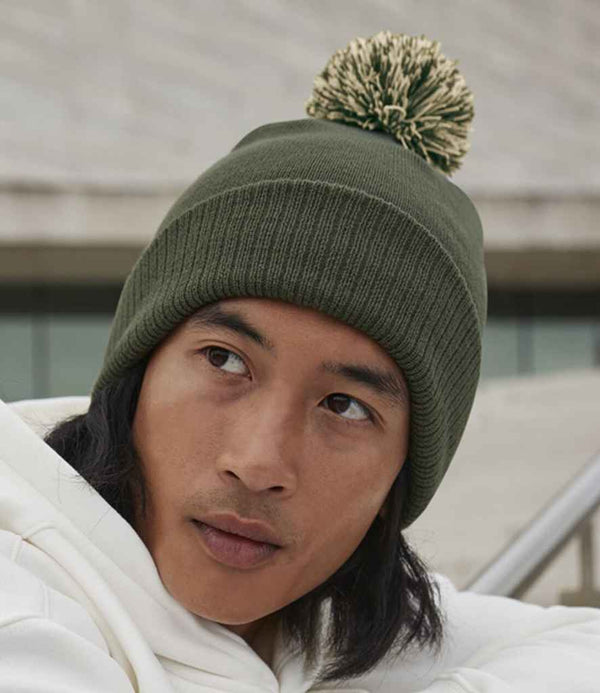 A person wearing a green Beechfield Snowstar® Beanie with a two-tone pompom and a white hoodie gazes off to the side. The outdoor background is blurred with visible concrete steps.