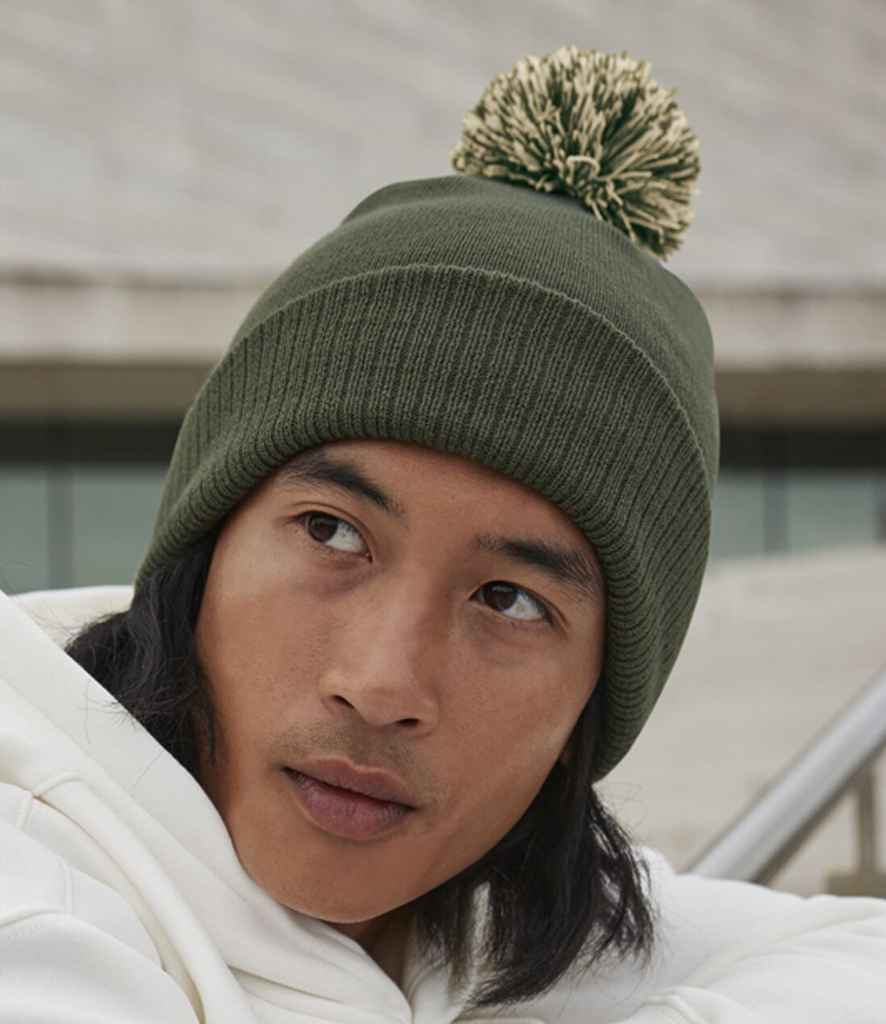 A person wearing a green Beechfield Snowstar® Beanie with a two-tone pompom and a white hoodie gazes off to the side. The outdoor background is blurred with visible concrete steps.